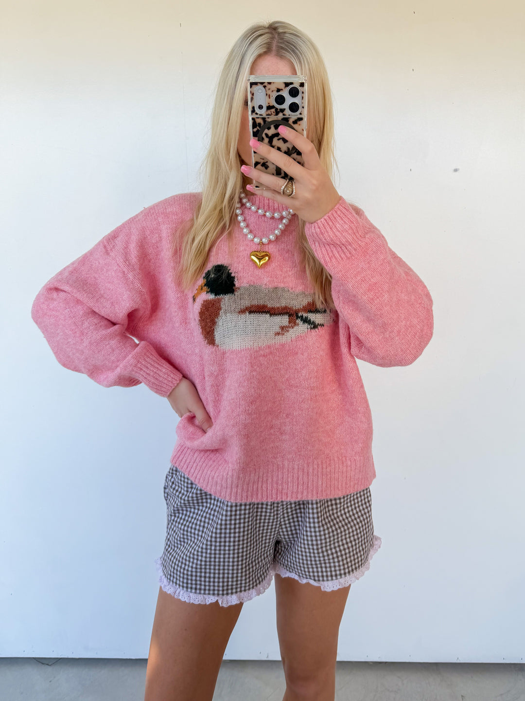 Person wearing a pink sweater with a graphic design and checkered shorts, taking a mirror selfie.