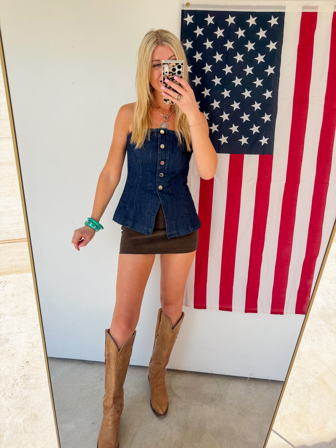 Person wearing a denim top and brown skirt with cowboy boots in front of an American flag.