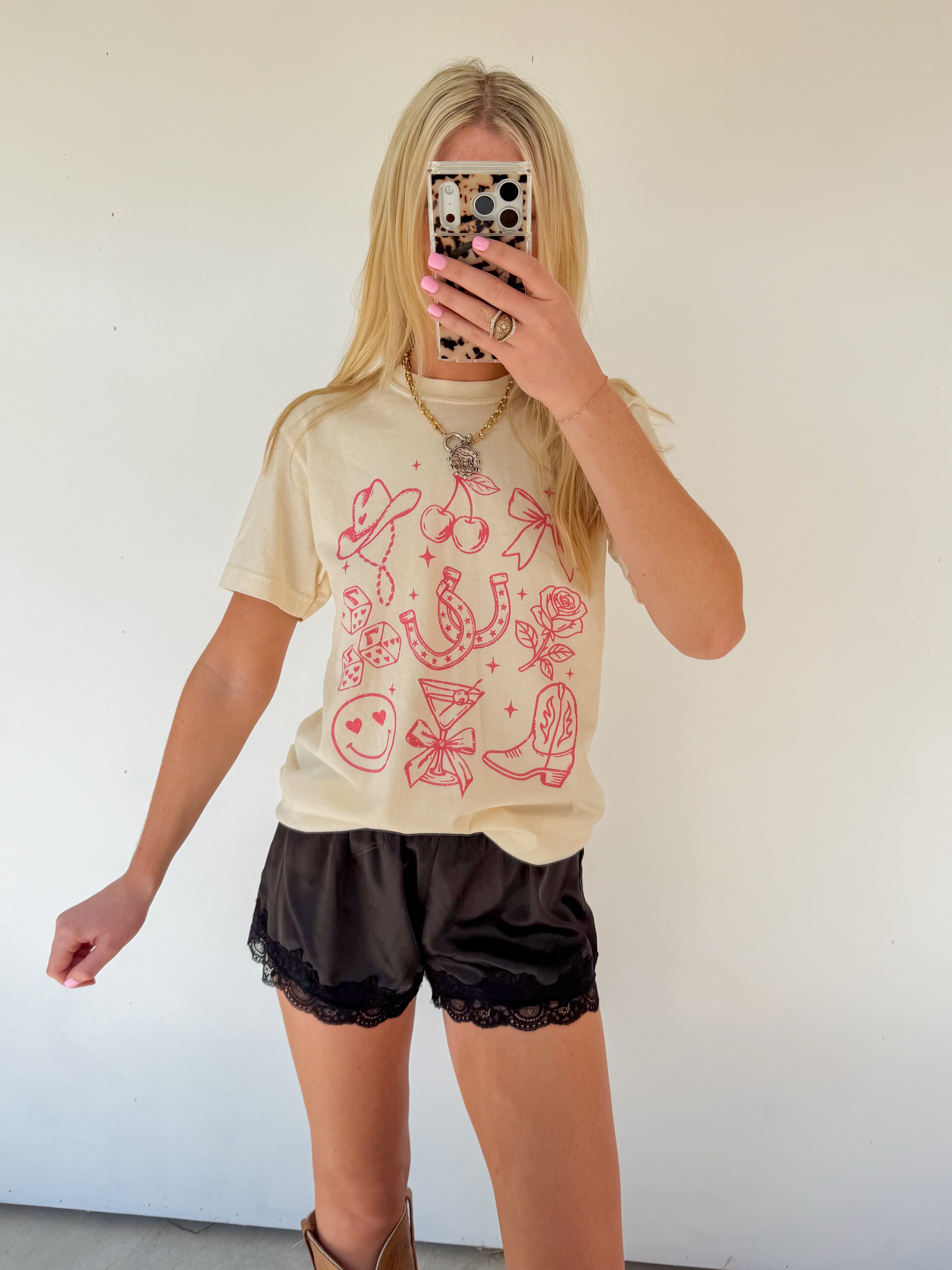 Person wearing a beige t-shirt with red graphics and black shorts, standing against a white wall.