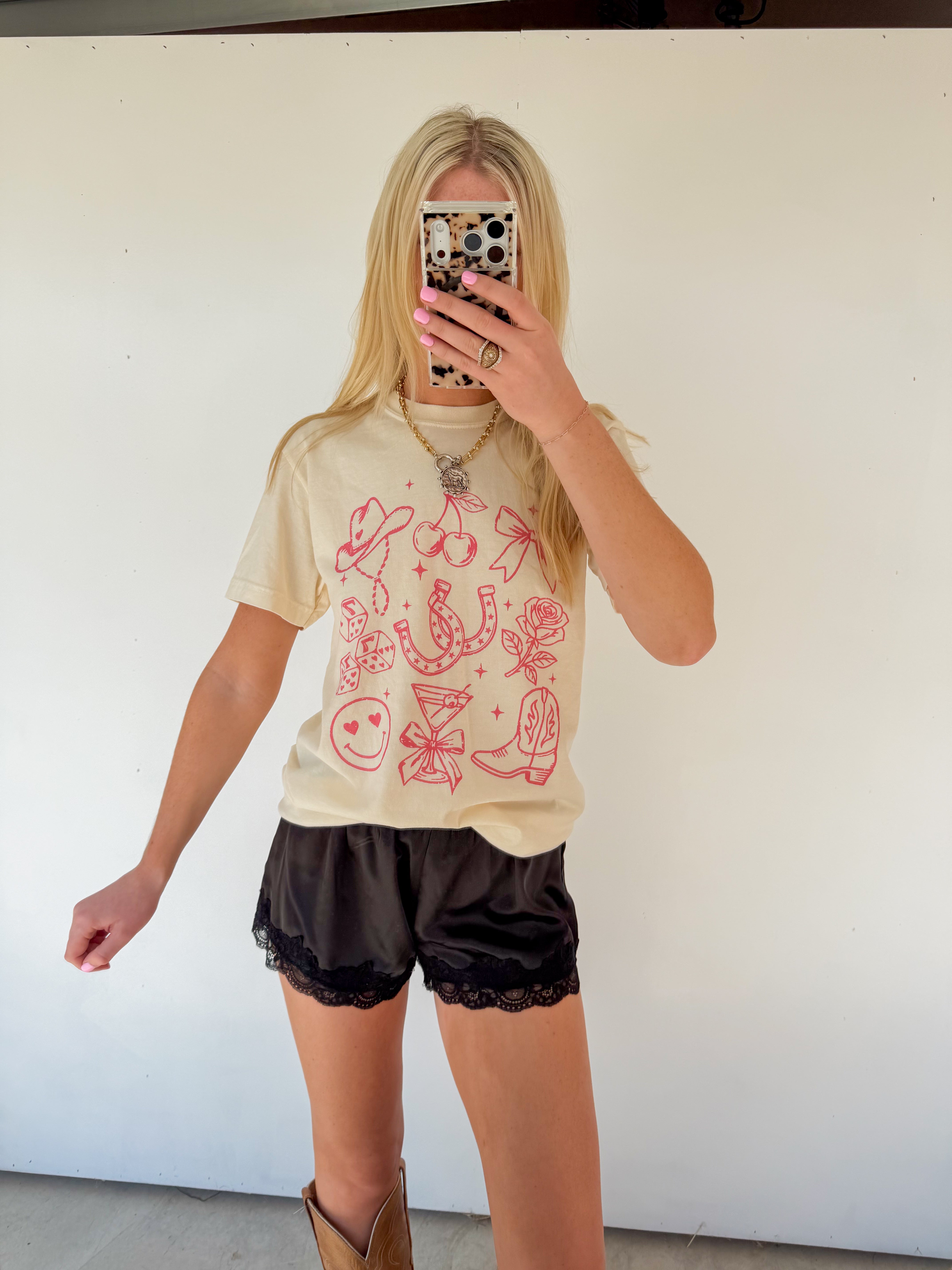 Person wearing a beige t-shirt with red graphics and black shorts, standing against a white wall.