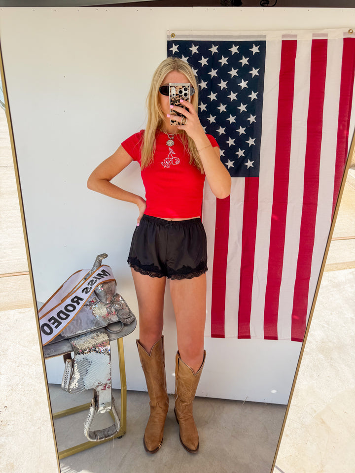 Person taking a mirror selfie wearing a red shirt, black shorts, and brown boots in front of an American flag.