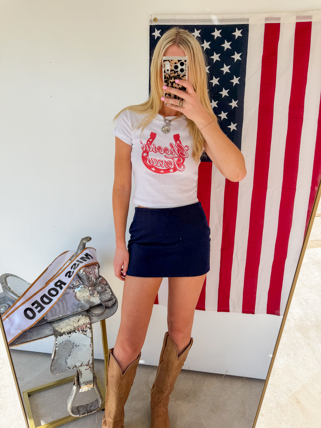 Person wearing a white t-shirt with red graphics and a navy skirt, standing in front of an American flag.