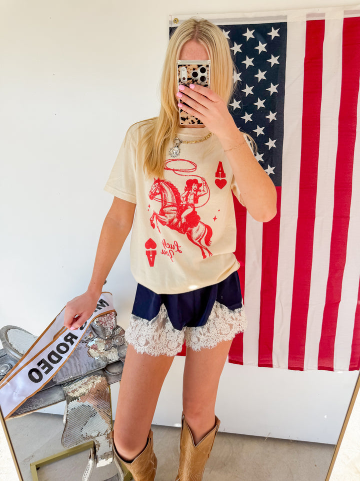 Person wearing a graphic t-shirt with a horse design, shorts, and boots in front of an American flag.