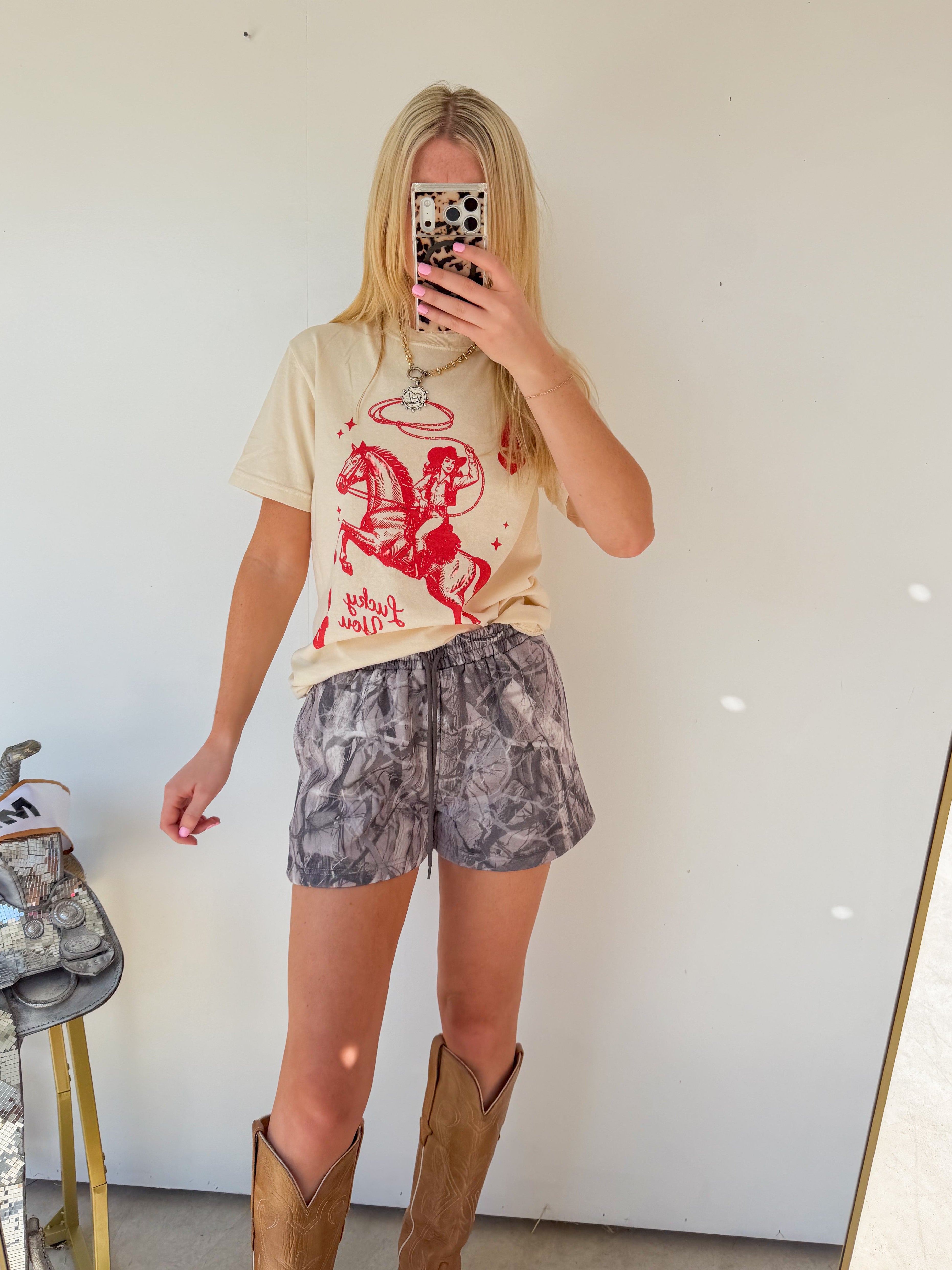 Person taking a mirror selfie wearing a graphic t-shirt, camouflage shorts, and brown boots.