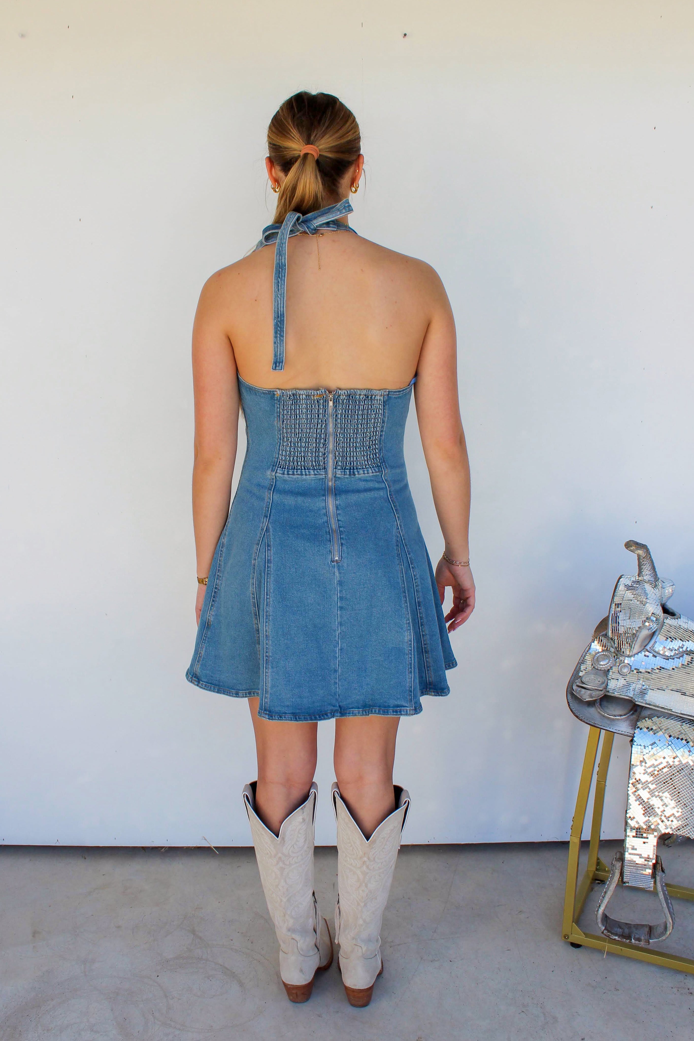 Woman wearing a blue denim dress and white boots standing against a white wall.