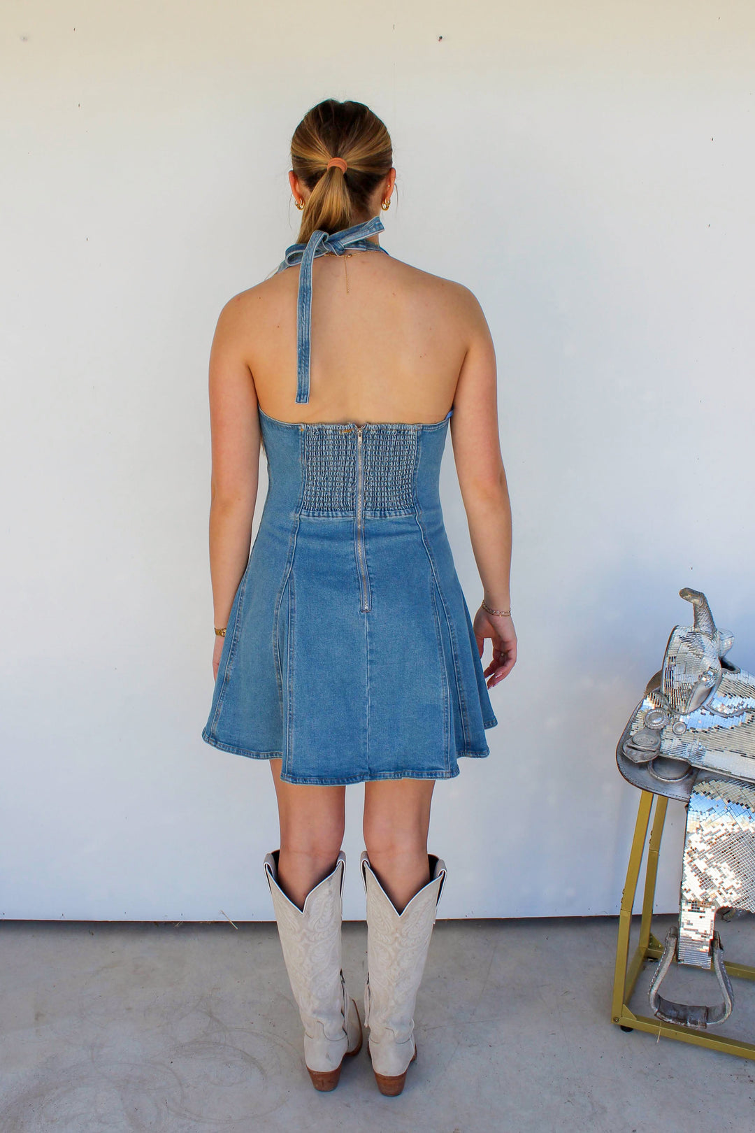 Woman wearing a blue denim dress and white boots standing against a white wall.