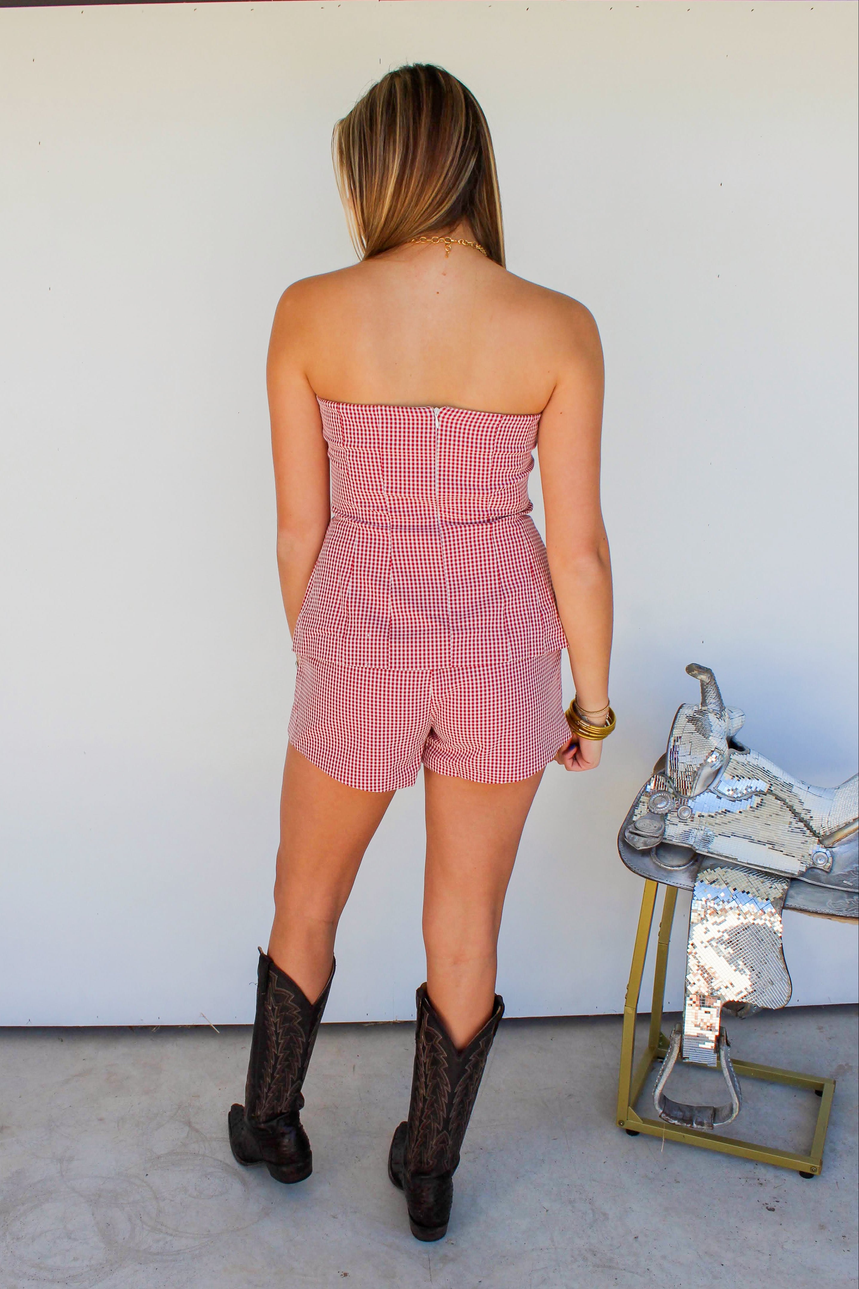 Woman wearing a pink checkered strapless romper and black cowboy boots standing in front of a white wall.