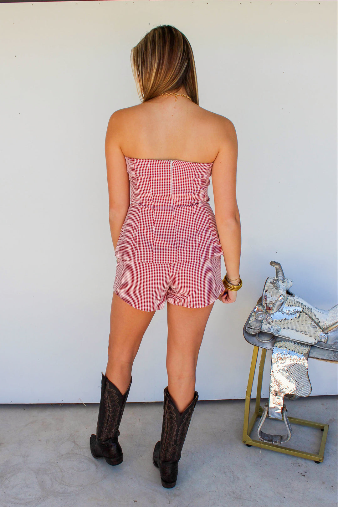 Woman wearing a pink checkered strapless romper and black cowboy boots standing in front of a white wall.