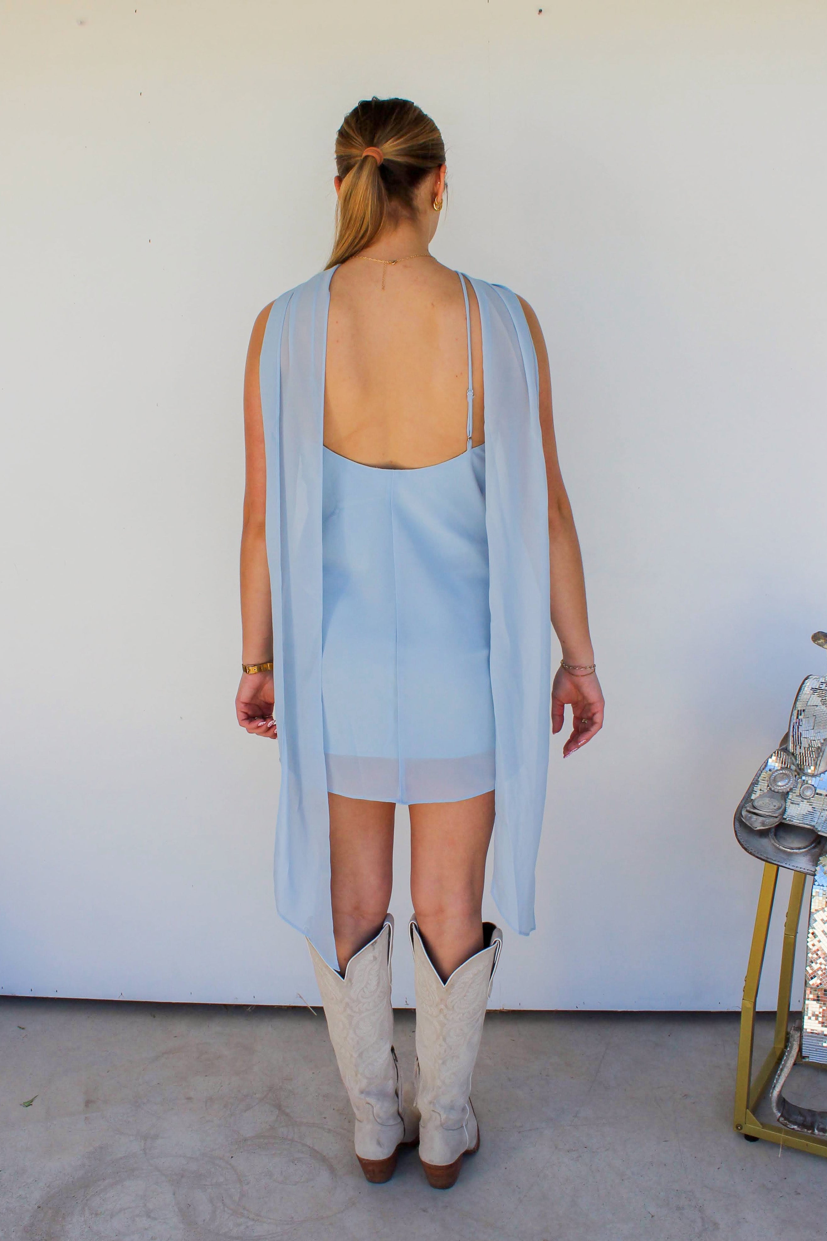 Person wearing a light blue dress with a long sheer overlay and white cowboy boots, standing in front of a white wall.