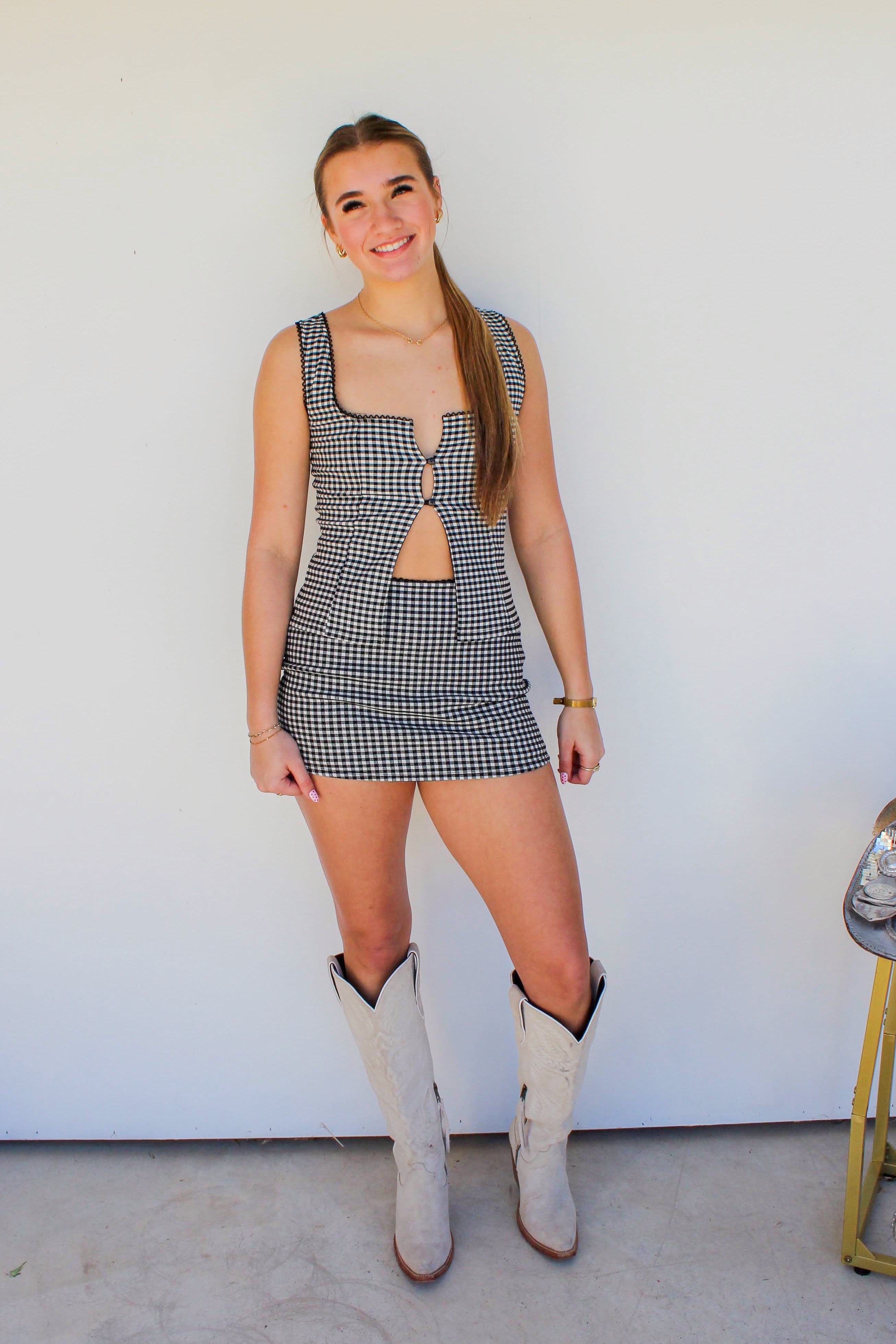 Woman in a checkered dress and cowboy boots standing in front of a white wall.