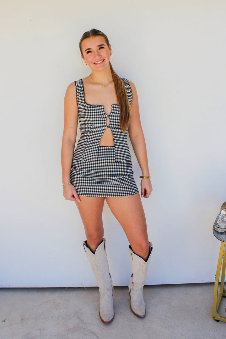 Woman in a checkered dress and cowboy boots standing in front of a white wall.