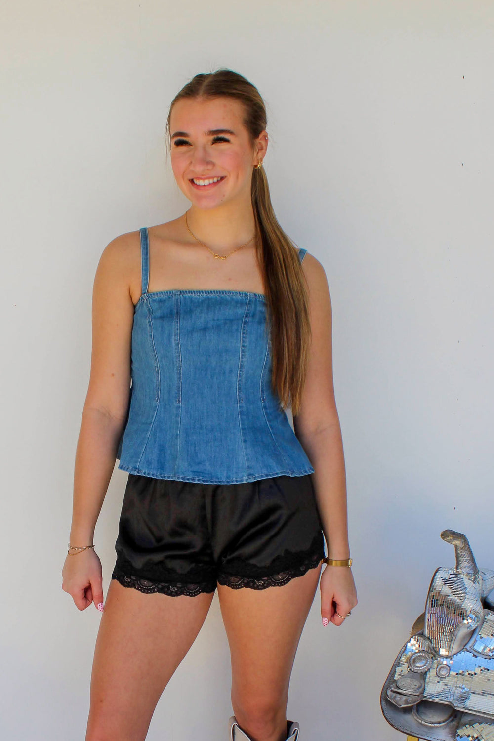 Woman wearing a denim top and black shorts with white boots standing against a white wall.