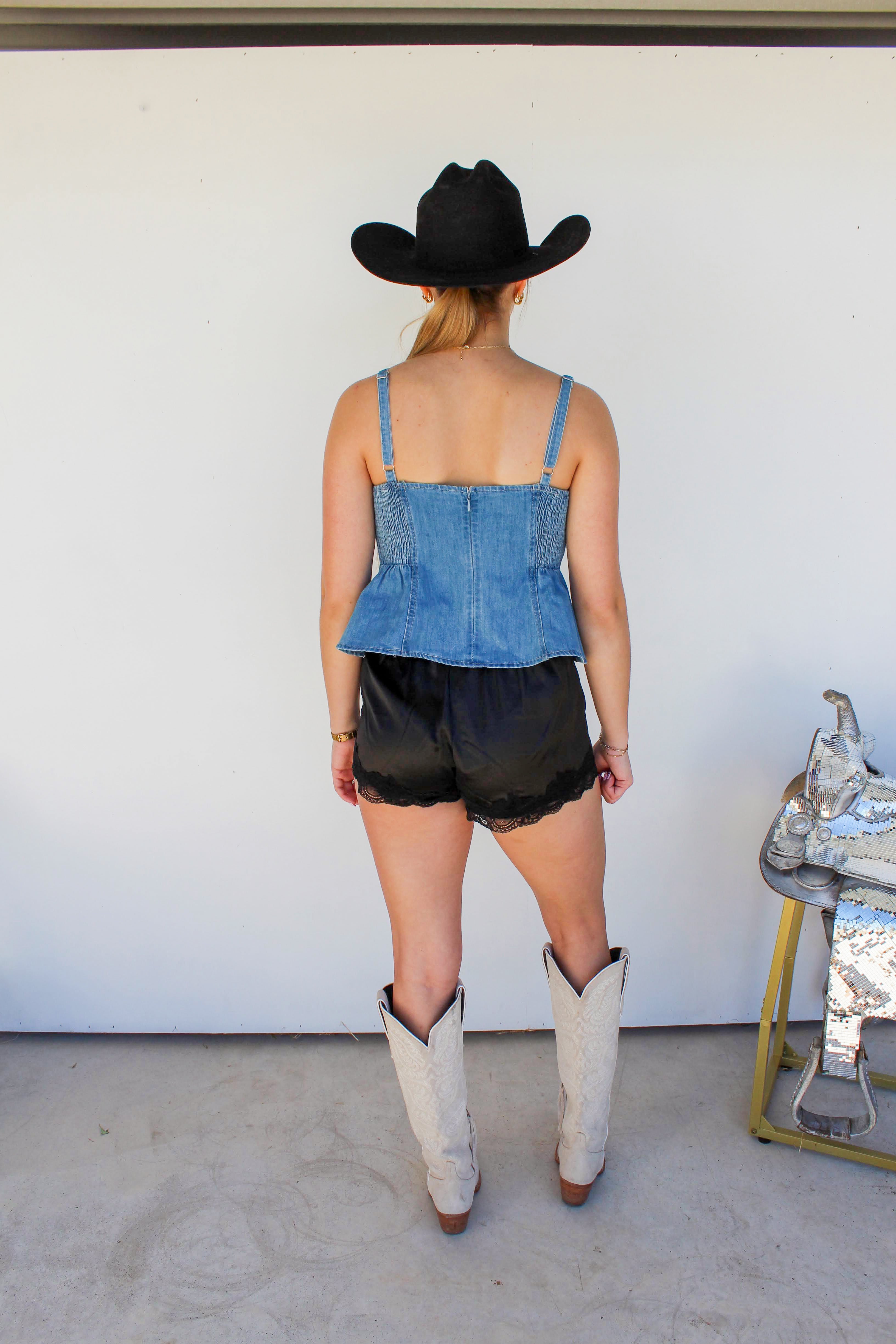 Person wearing a denim top, black shorts, and white cowboy boots with a black cowboy hat, standing in front of a white wall.