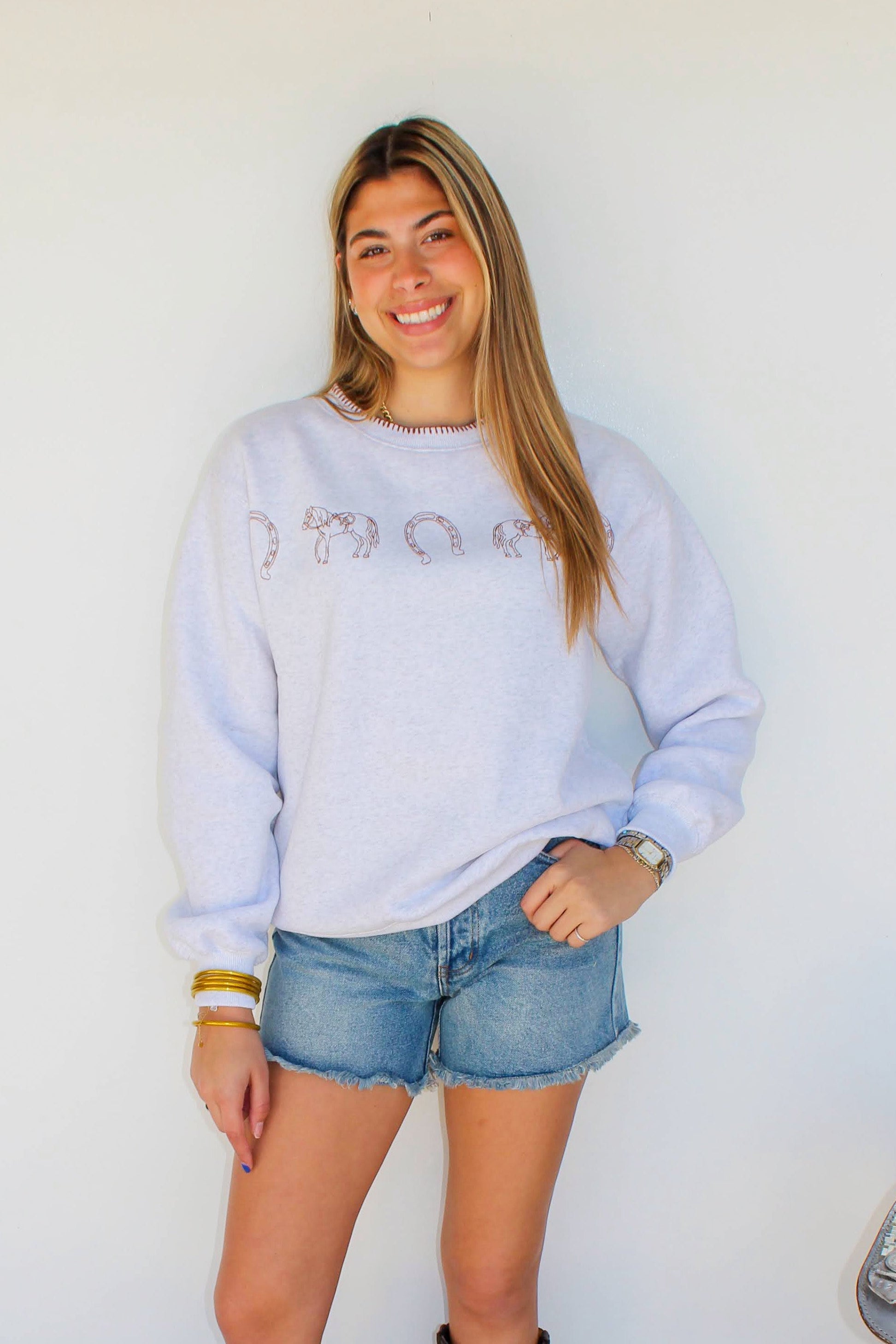 Woman wearing a light blue sweatshirt, denim shorts, and dark boots standing in front of a white wall.