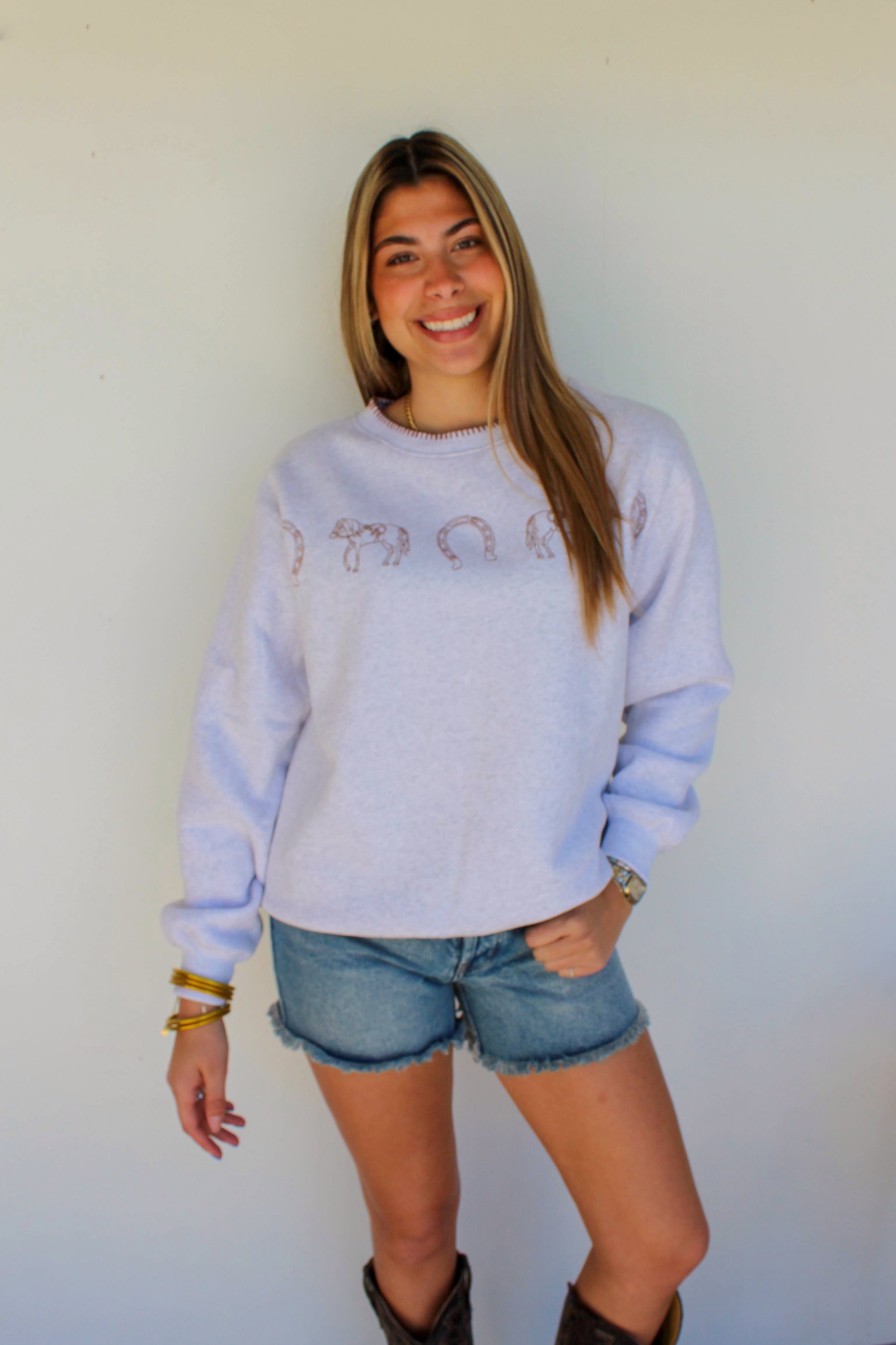 Woman wearing a light blue sweatshirt with text, denim shorts, and brown boots against a white wall.
