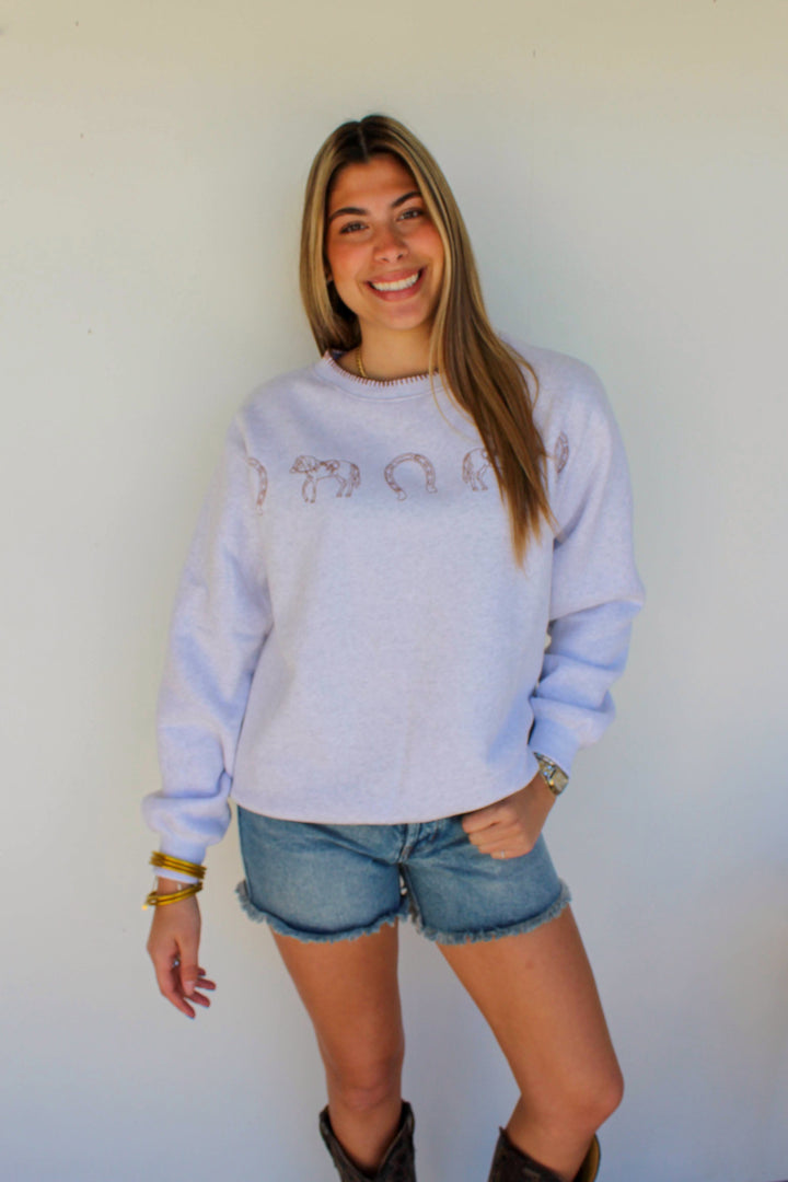 Woman wearing a light blue sweatshirt with text, denim shorts, and brown boots against a white wall.