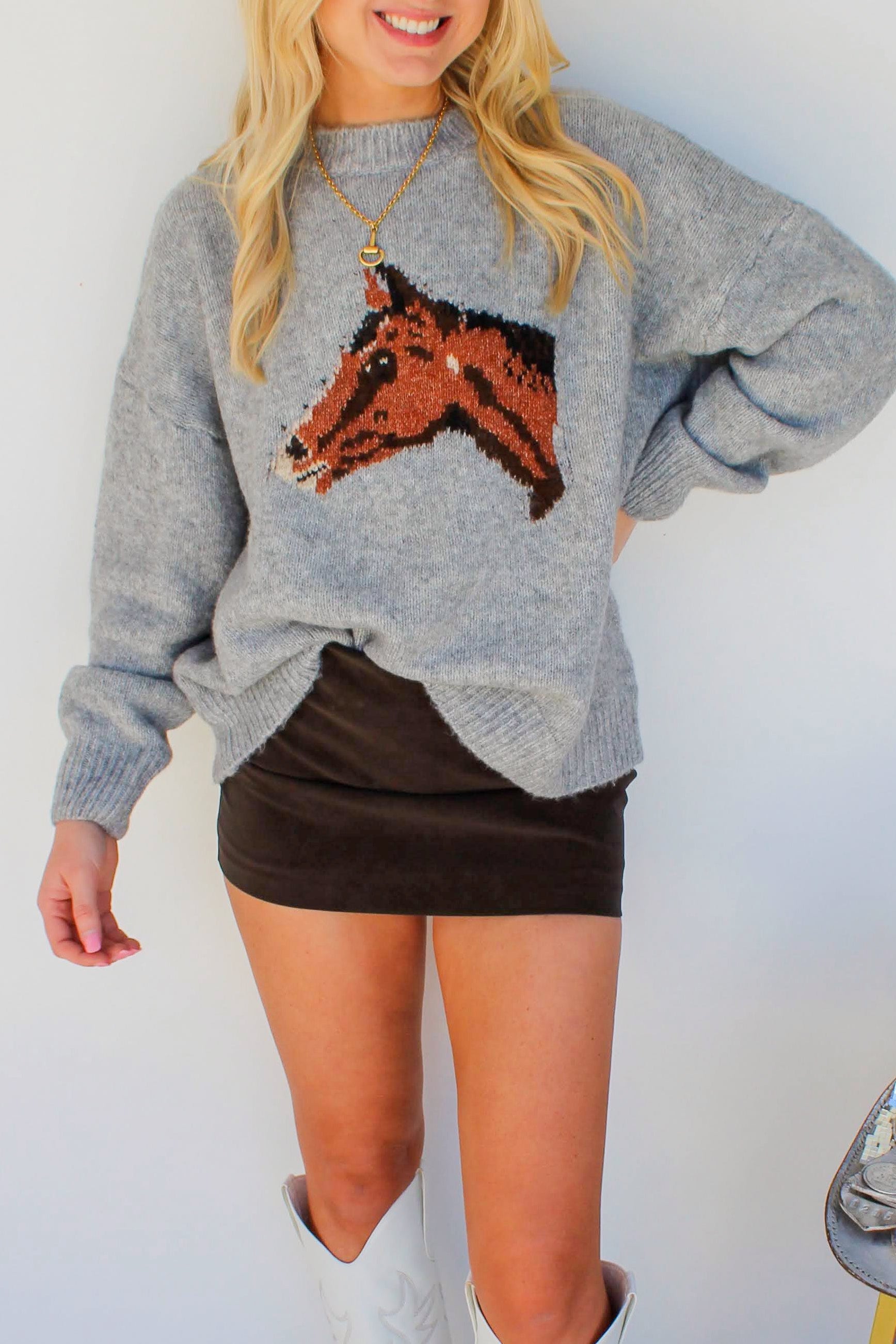 Woman wearing a gray sweater with a horse design, black skirt, and white boots standing next to a silver saddle.