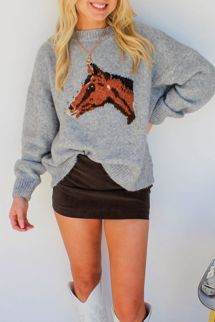 Woman wearing a gray sweater with a horse design, black skirt, and white boots standing next to a silver saddle.