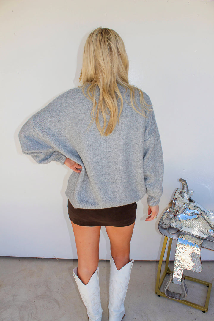 Person wearing a gray sweater and white boots standing in front of a white wall with a mirror on a stand.