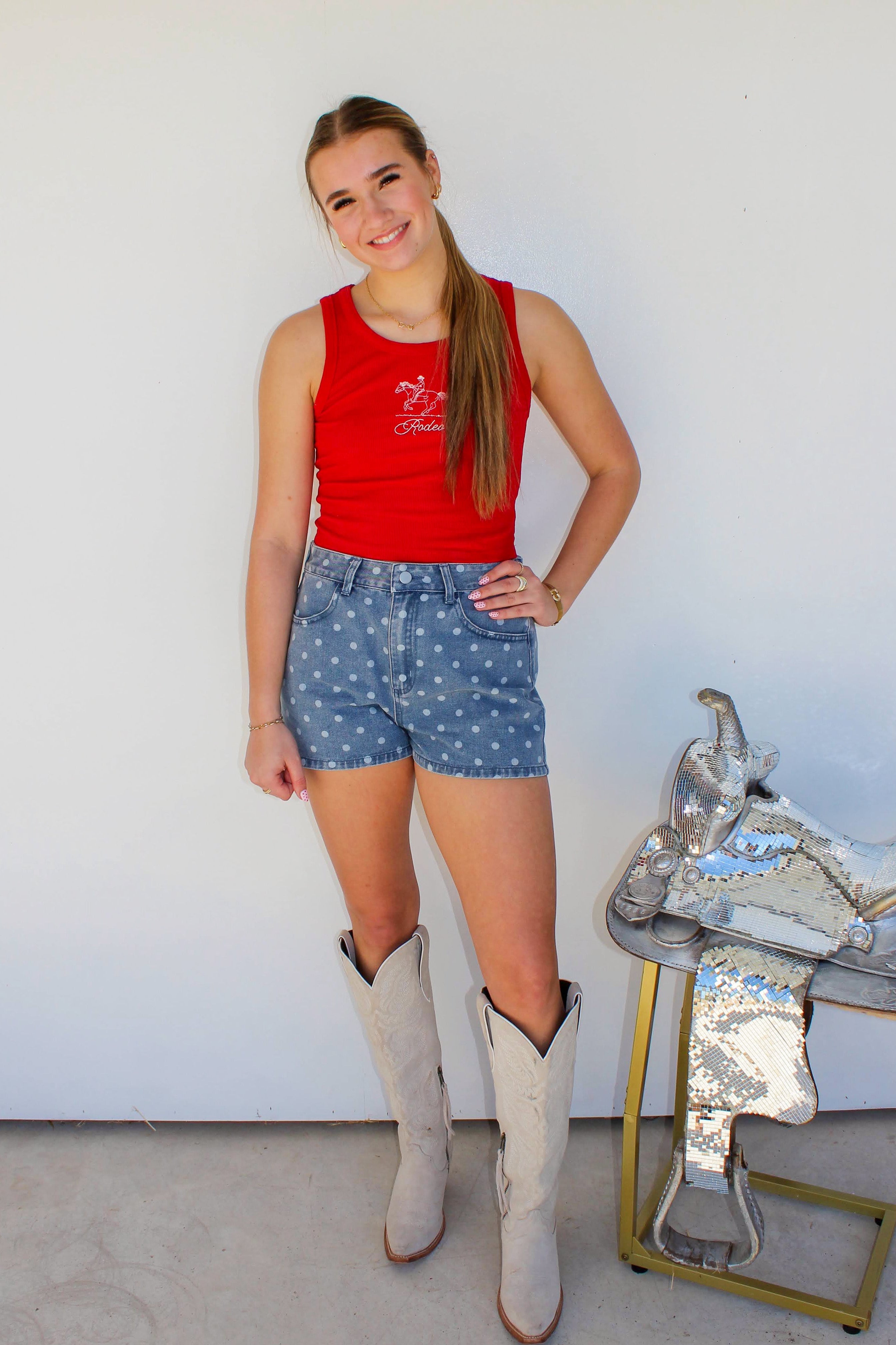 Woman in red tank top and blue shorts standing next to a silver horse saddle against a white wall.