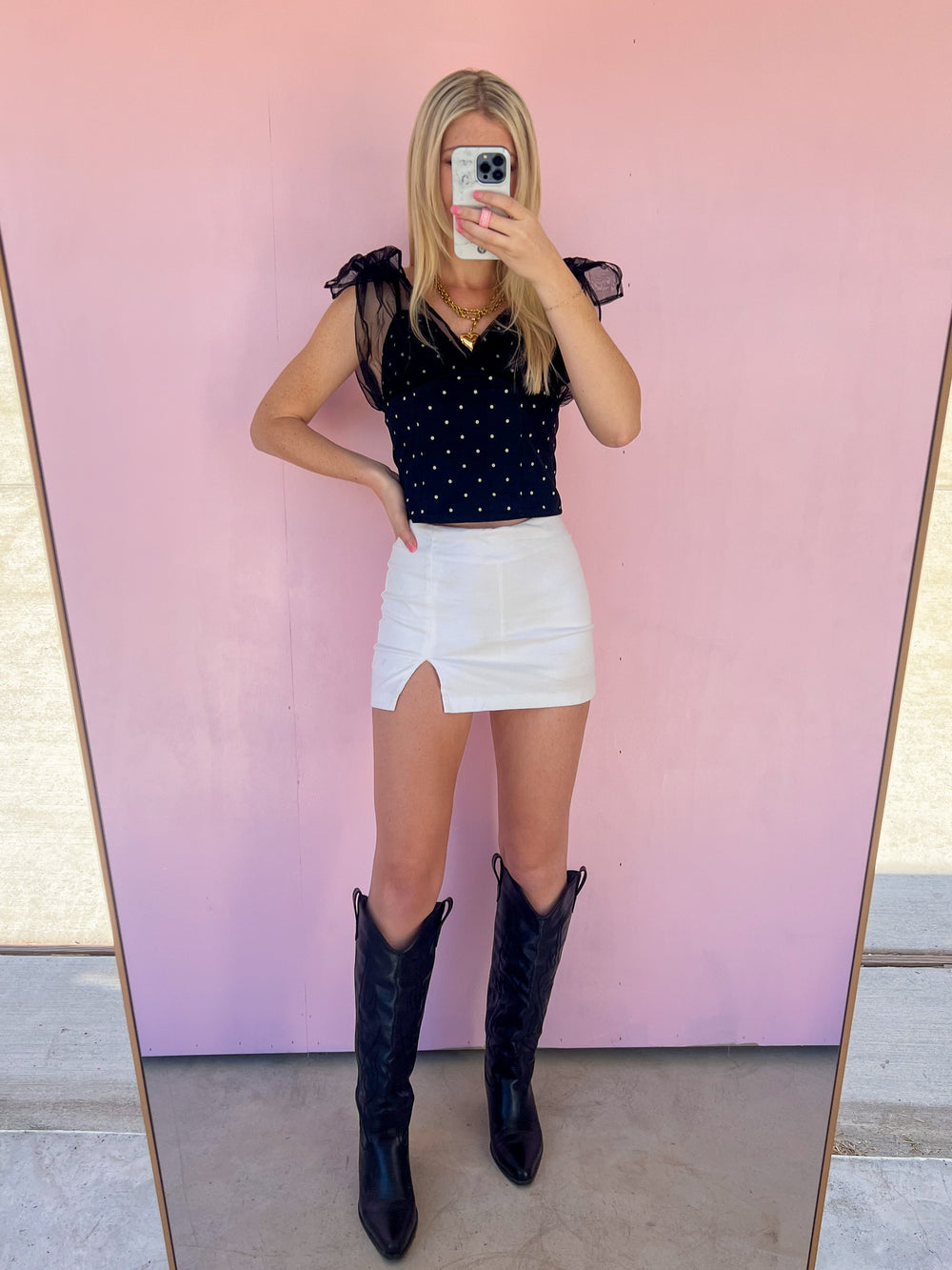 Person wearing a black polka dot top, white skirt, and black cowboy boots in front of a pink wall.