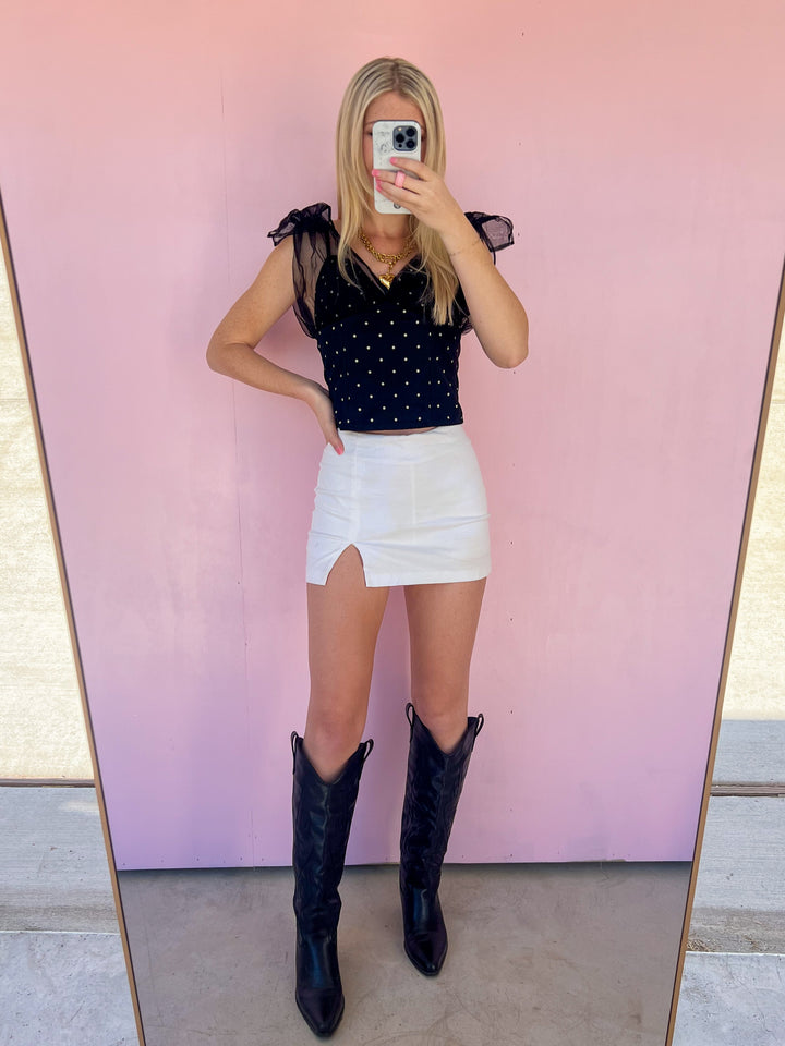 Person wearing a black polka dot top, white skirt, and black cowboy boots in front of a pink wall.