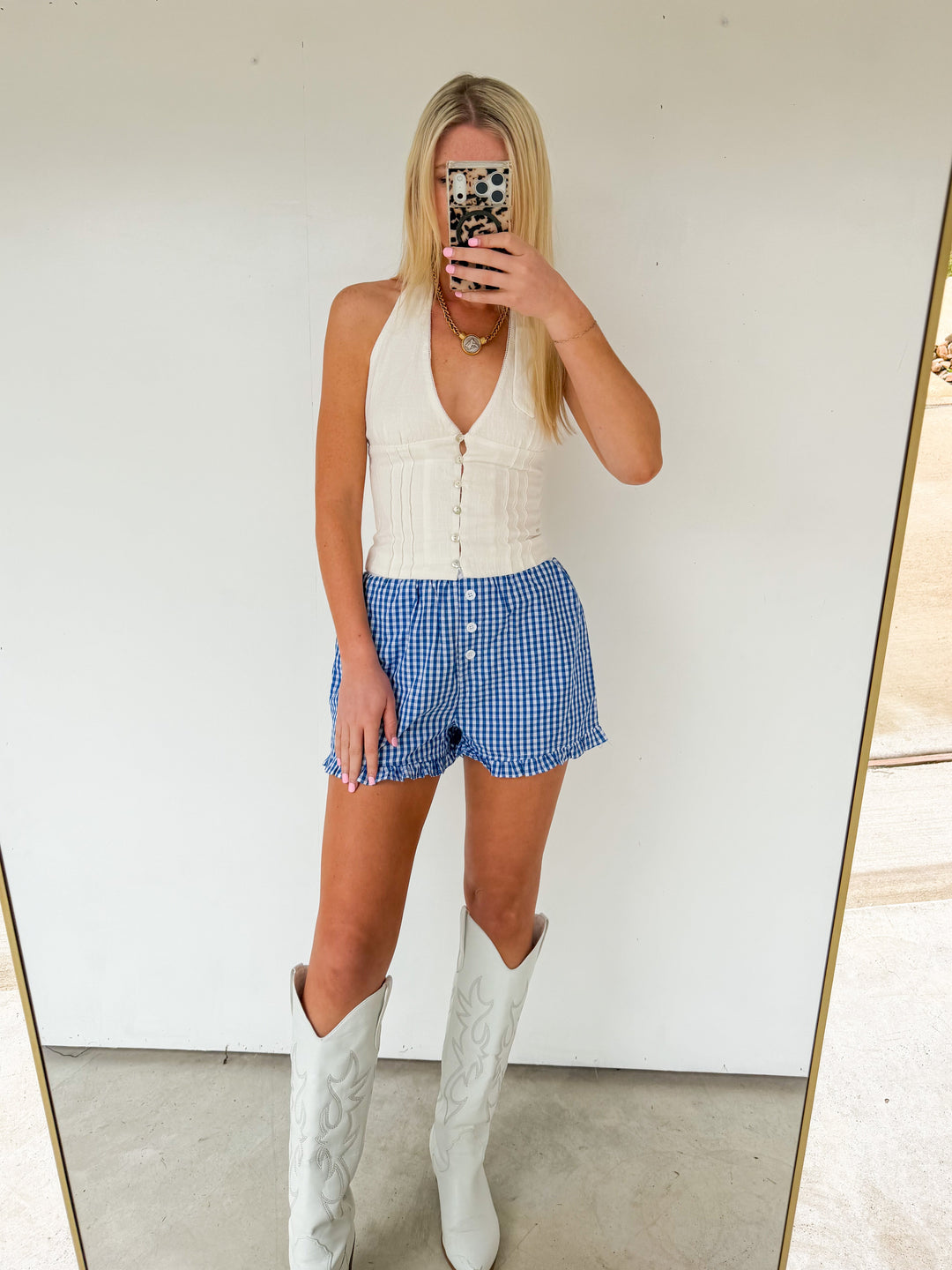Person wearing a white sleeveless top, blue checkered shorts, and white knee-high boots in front of a mirror.